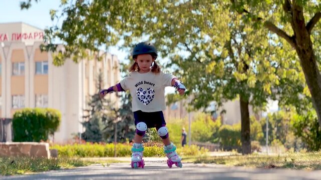 Little girl wearing a helmet and protective gear roller skating along a park path. Children's sports, active recreation, movement and happy carefree childhood concept