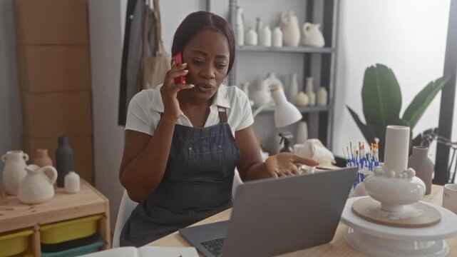 Woman potter in apron holding phone to ear while checking laptop, clay vases, brushes and packaging on the studio table; concerned entrepreneur.