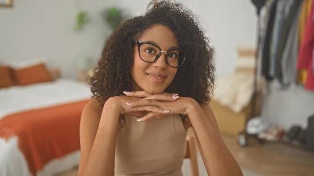 Woman smiling with hands clasped under chin, seated in a bedroom near bed and clothing rack; calm focus.