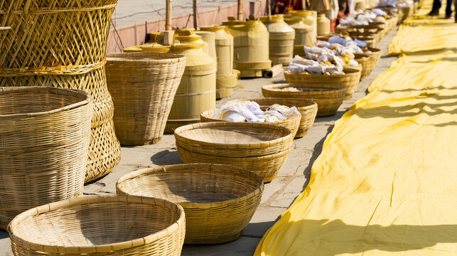 Traditional Hindu Antyesti ritual preparations with woven bamboo baskets, ceremonial containers and wrapped offerings arranged in a long row beside a yellow cloth pathway under bri