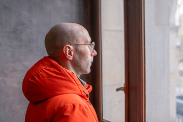 Bald middle-aged man in orange winter jacket and glasses looking window, thoughtful male profile in bright outerwear indoors, urban lifestyle and solitude, Interior portrait, historical architecture © kittyfly