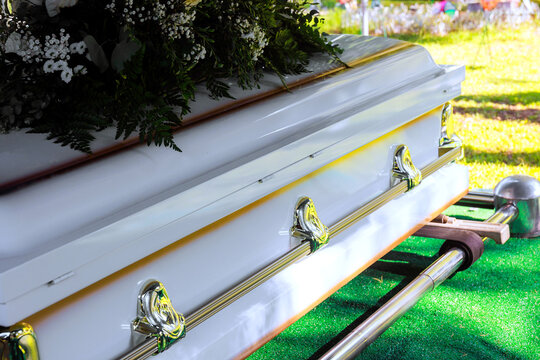 Coffin sits on green grass at burial site, flowers decorations surround cemetery area, family stands nearby.