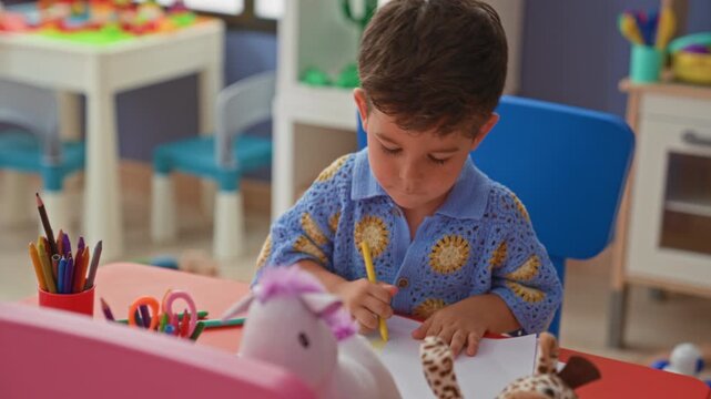 Boy cutting paper with orange safety scissors, hand positioned on paper at a preschool table with crayons and toys visible; concentration.