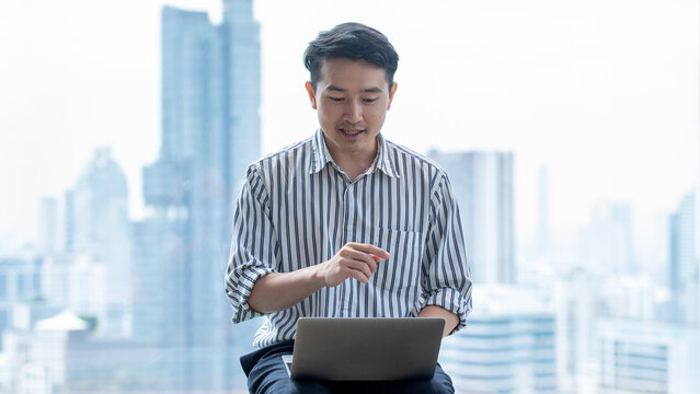 Asian Businessman Using Laptop in Modern City Office with Urban Skyline Background