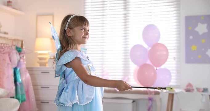 Smiling little girl in fairy costume with wings and headband playing with magic wand at home. Camera moving forward and backwards
