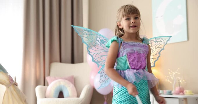 Smiling little girl in beautiful costume with wings waving magic wand at home