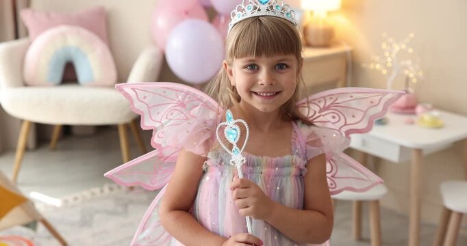 Smiling little girl in fairy costume with wings and magic wand at home. Camera moving forward and backwards