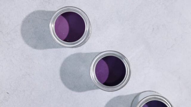 Cans with paint in shades of purple and brush on light grey table, flat lay. Camera moving up