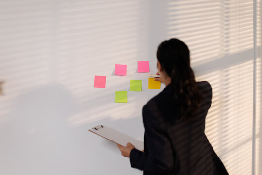 Professional woman organizing ideas on a white wall with colorful sticky notes, planning business strategy, and brainstorming project tasks in an office environment