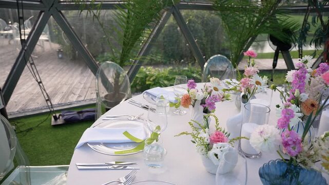 Beautifully arranged round table with pastel flowers, clear chairs, and modern dinnerware in a glass dome, surrounded by greenery and soft natural daylight, creating a festive mood.
