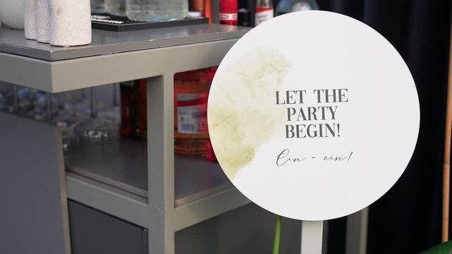 Detailed close-up shot of a round sign reading 'Let the Party Begin!' beside a bar cart with assorted bottles, glassware, and festive decor, bright daylight fills the scene.