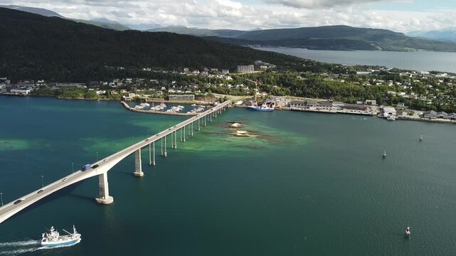 Aerial drone footage pushes forward toward the Gisund Bridge in Finnsnes, Norway, as a fishing boat passes beneath it and a truck and cars cross above