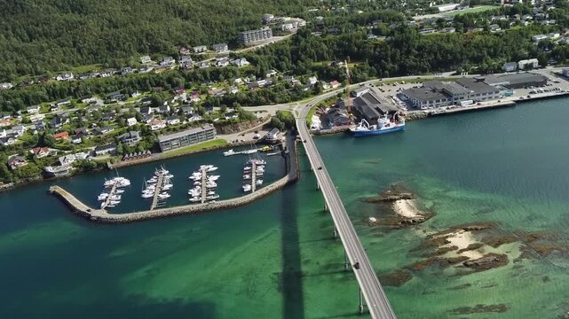 Cars cross the Gisund Bridge above a marina of docked boats and a cargo ship at the Finnsnes harbor, with sandbanks visible through turquoise water, aerial view