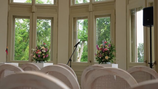 Beautifully arranged empty wedding ceremony venue featuring tall windows, soft daylight, rows of white chairs, floral displays, and a microphone stand. Serene, airy ambiance, no people present.