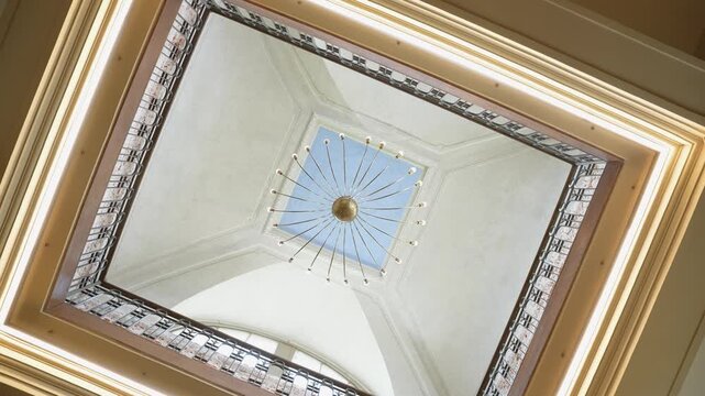 Dynamic camera movement captures an ornate square atrium ceiling with skylight and radial chandelier, framed by intricate railings and warm interior lighting in a stylish architectural space.