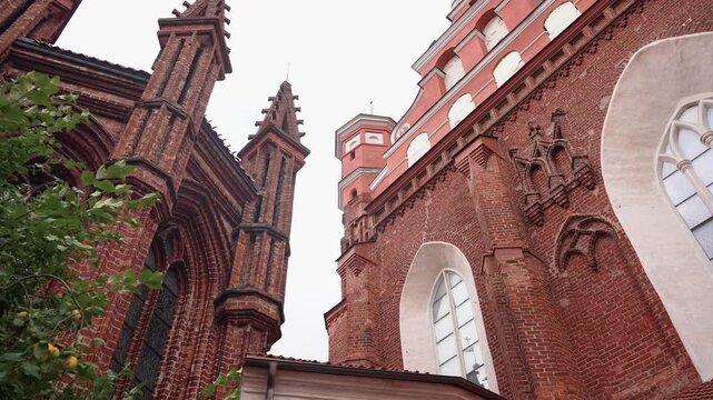 Low angle shot reveals intricate red brick Gothic architecture of the Church of St. Francis and St. Bernardino in Vilnius, with pointed spires, lush greenery, and soft daylight.