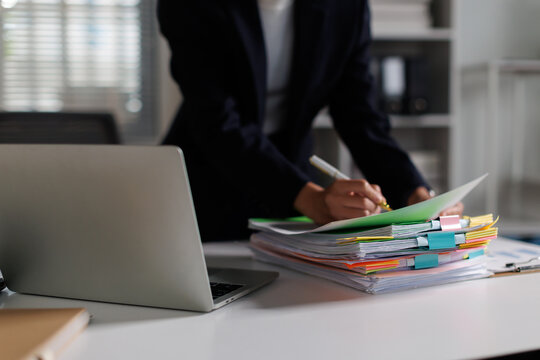 Businesswoman standing, reviewing financial reports and paperwork, signing documents on a cluttered office desk with a laptop, managing important corporate data and tasks