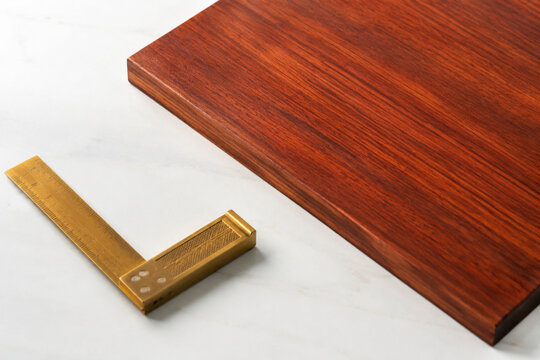 Polished padauk wood slab with brass carpenter's square, bright studio surface, wide crop
