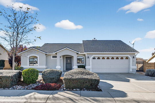 Modern suburban house with white facade arched windows gray roof manicured front yard trees sky day