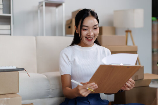 Asian small business owner managing online store inventory, checking customer orders on a clipboard, and packing cardboard boxes for e commerce startup delivery