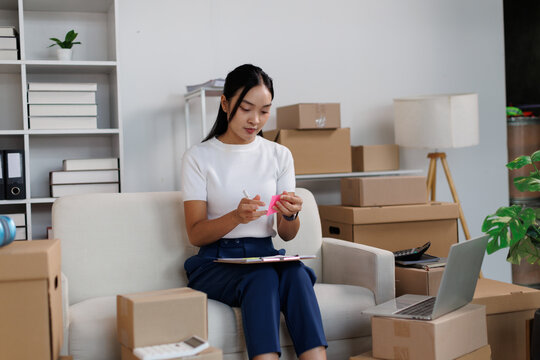 Asian woman concentrating on writing a note for a package, surrounded by cardboard boxes and a laptop in a modern home office, symbolizing effective management of an online retail business