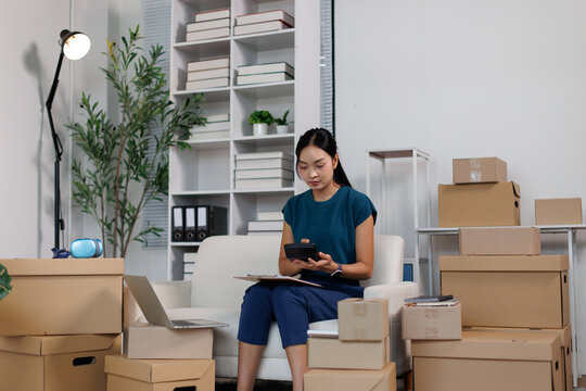Young asian woman managing her online small business from home, calculating orders and preparing packages for e commerce shipping in a startup office