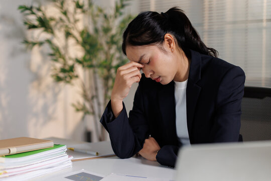 Overworked young asian businesswoman experiencing severe headache and eye strain at her desk, feeling stressed and exhausted from long hours and high pressure in a corporate office