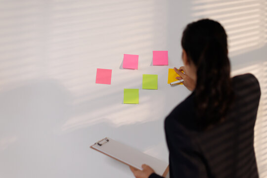 Businesswoman brainstorming ideas on a whiteboard, writing notes on colorful sticky papers, developing a new business strategy, planning startup project goals, organizing tasks in an office