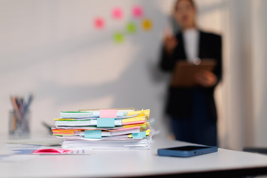 Stack of various reports and paperwork with colorful tabs and clips on an office table, showing a busy workflow and administrative tasks with a blurred person in a business setting