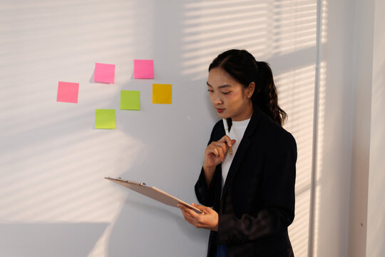 Businesswoman standing in office thinking and analyzing data on clipboard, brainstorming new project ideas with colorful sticky notes on wall