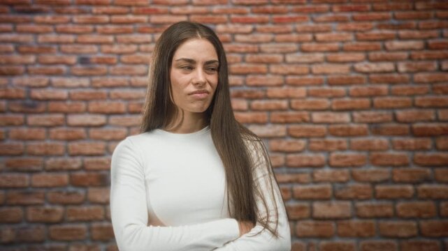 Young hispanic woman with arms crossed, long hair over shoulder, subtle smirk and visible forearm in studio brick wall setting; confidence.