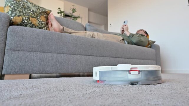 Smart vacuum cleaner cleans the carpet while man rests on the sofa. A smart home, modern technologies,housekeeping concept