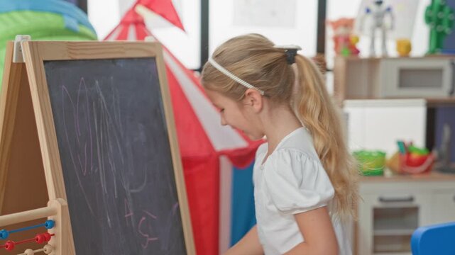 Blonde girl erasing a chalkboard with hand while holding sponge in a colorful playroom building; concentration childhood creativity.