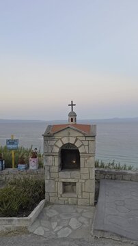 A low angle shot shows the cross atop a small stone shrine silhouetted against the sky and calm sea in Afytos Chalkidiki Greece