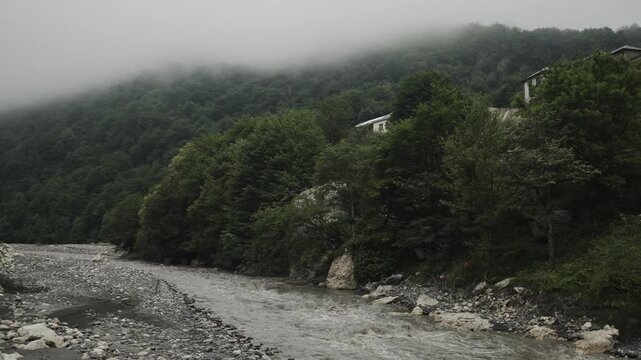 A rocky river flows below a forested hillside covered in low cloud in Gabala in daylight.
