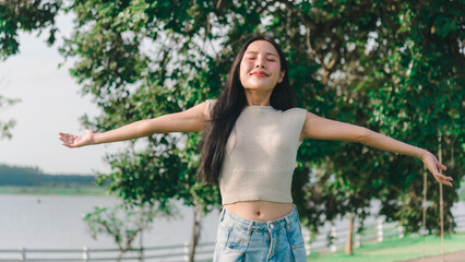Smiling young woman expressing gratitude outdoors during sunny day portrait, feeling happiness and peace with arms open wide in nature park environment under bright natural sunlight illumination