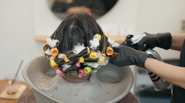 Hairdresser wearing black gloves sprays neutralizing solution between perm rollers on back of woman's head