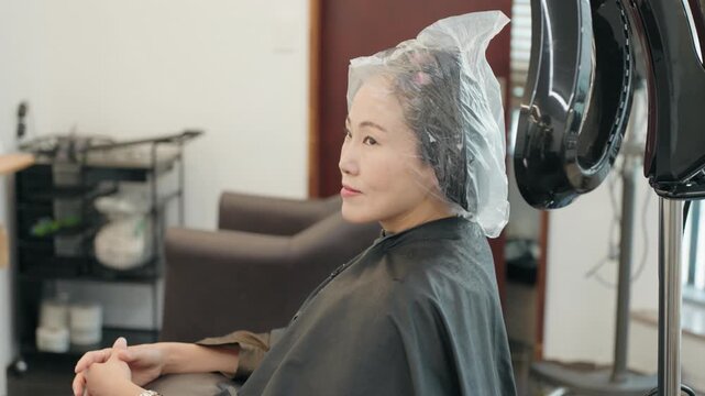 Camera moves around woman wearing plastic cap while sitting under steaming perm machine during salon treatment