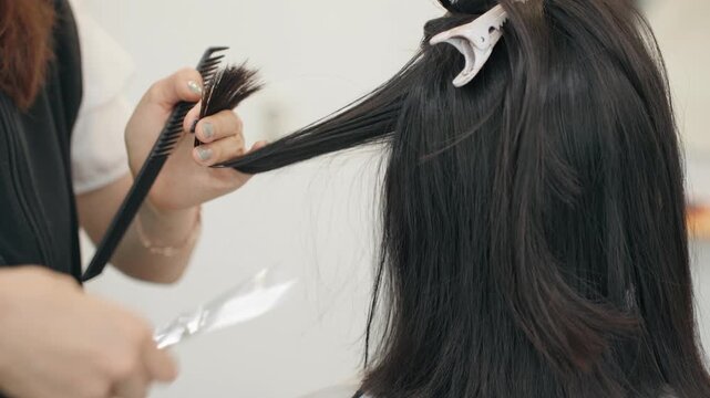 Hairstylist combs woman's long dark hair section with clip while rotating scissors onto pinky finger between cuts