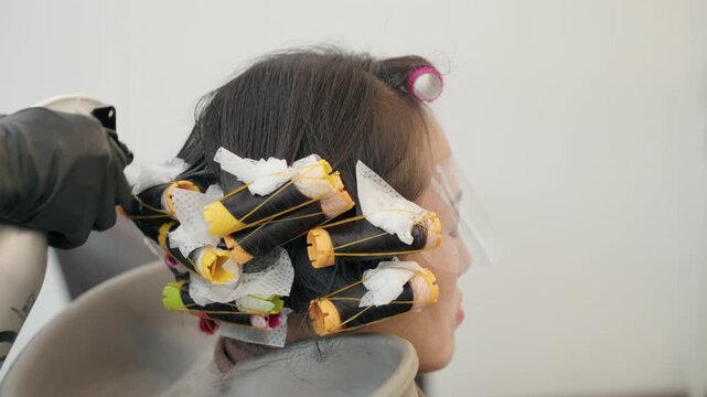 Hairdresser wearing black gloves adjusts perm rollers wrapped in tissue paper on woman's hair with face shield