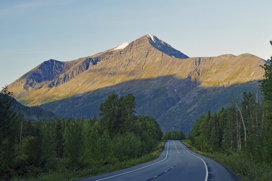 Traffic-free road leads as grades towards rugged mountains illuminated by the morning sun, Richardson Highway, Alaska, USA