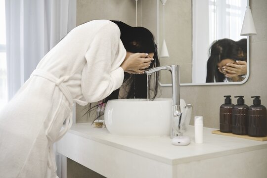 A woman in a white bathrobe leans over a stylish sink in a contemporary bathroom, splashing water on her face