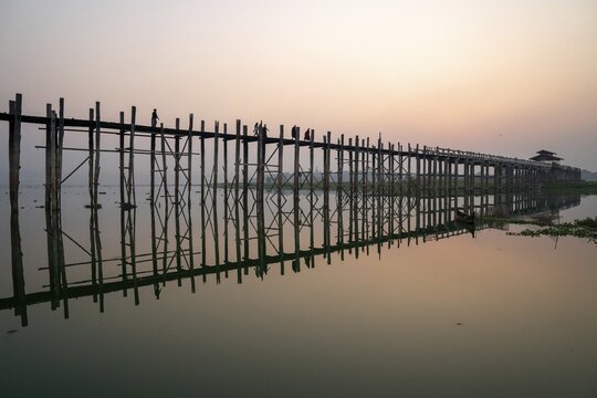 Buddhist monks walk across the U-leg bridge in red robes as the sun rises, Mandalay, Myanmar