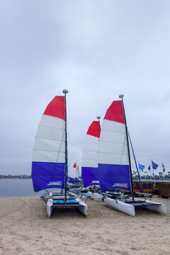 Small catamaran sailboats with red white and blue sails parked on a sandy Mission Bay beach under a cloudy sky, San Diego, CA