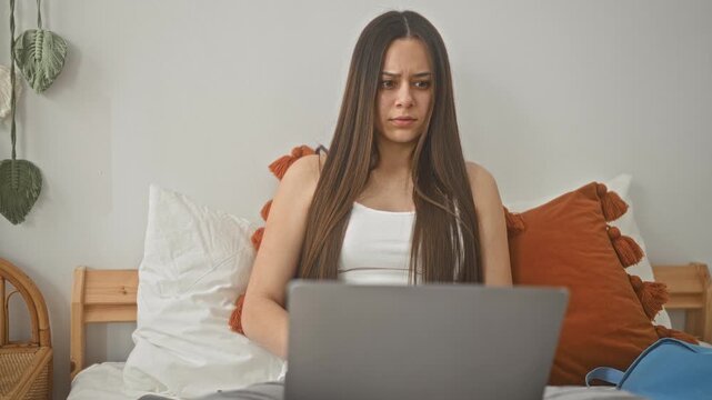 Woman on bed using laptop with hand covering face in bedroom, studying and squinting at screen; stress study pressure.