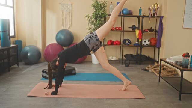 Woman on yoga mat performing three legged downward dog pose, raising left leg with hands grounded in studio among fitness balls and shelf gear; serenity.