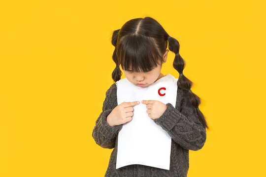 Little schoolgirl looking at her test paper with a bad grade C and feeling sad.