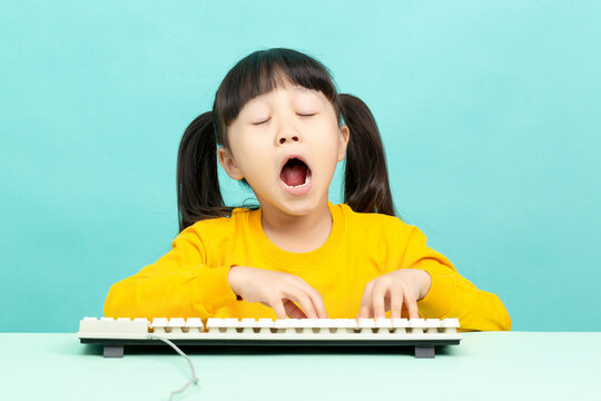 Tired little girl yawning with her eyes closed while typing on a computer keyboard.