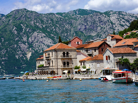 Traditional red-roofed stone villas and moored excursion boats line the scenic waterfront of Perast along the Bay of Kotor beneath towering limestone mountains in Montenegro