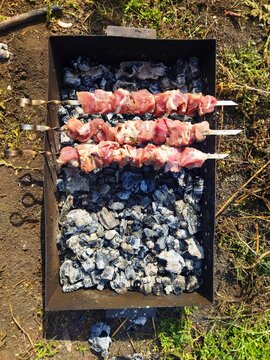 meat on the grill,Top-down view of three metal skewers loaded with raw pork shish kebab (shashlik) pieces, placed on a black metal barbecue grill over glowing and ashen charcoal in an outdoor garden 